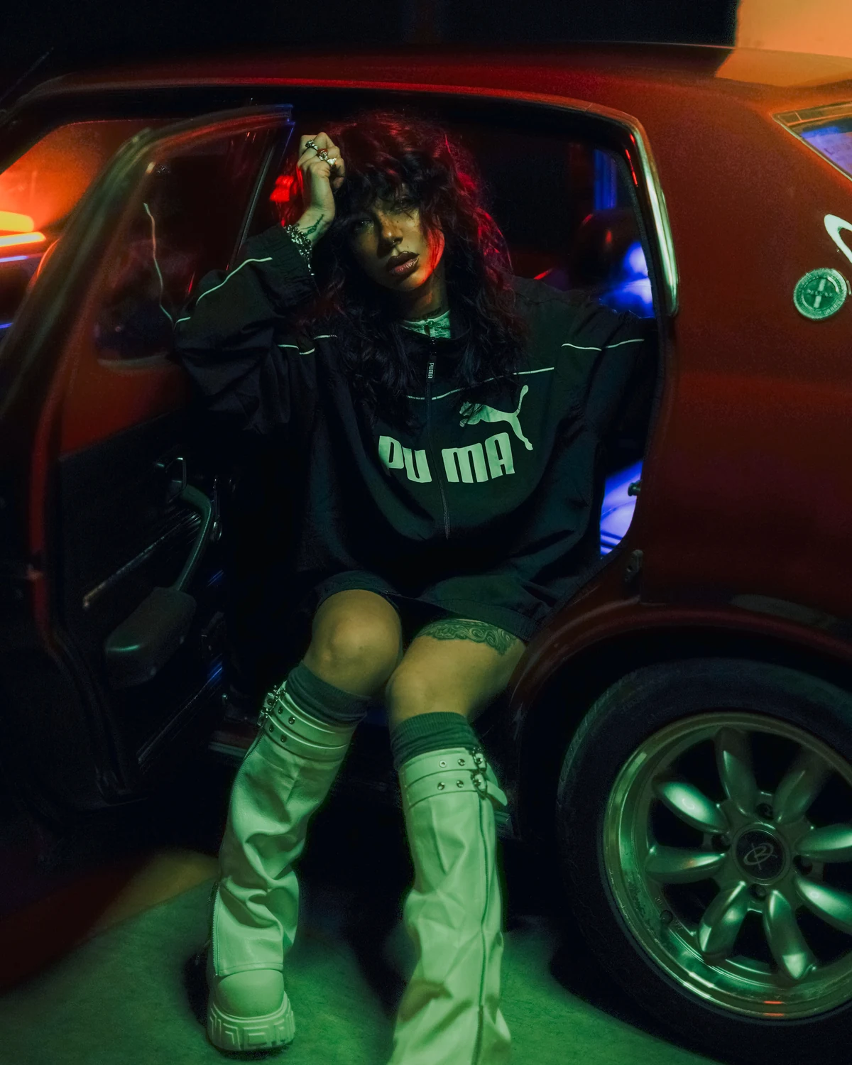 Anastasiya Ty seated beside a car in a green and orange night scene